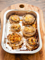 Cooked high quality stuffed mushrooms in a foil tray on wooden board and table. Fine starter dish with premium ingredients .