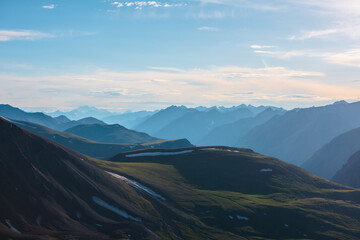 Aerial top view to big green hills and large rocky mountains silhouettes in bright sun under clouds in blue sky. Sunlit high snowy mountain range far away. Scenic layered alpine landscape at sunset.