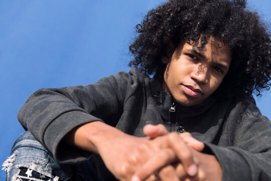 close up portrait of a thoughtful curly haired black male teenager in informal wear looking at camera while sitting against blue background, concept of young lifestyle