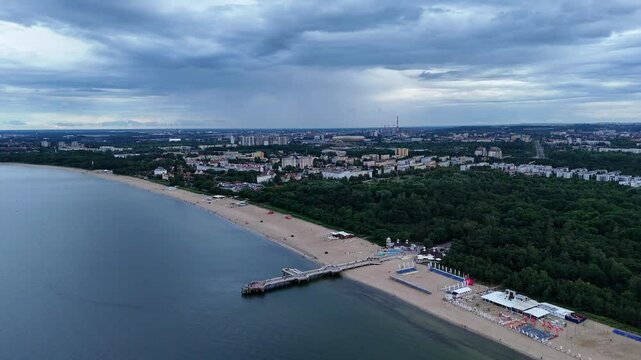 Gdansk beach stretches along the coast with a prominent pier extending into the sea. The cityscape in the background features a mix of urban and green areas under a cloudy sky drone revealing shot
