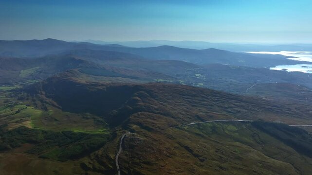 Caha Pass, County Cork, Ireland, September 2024. Drone tracks right in a panoramic high angle overview of a winding road crossing the rugged mountain ridge and descending to the North Atlantic Ocean.