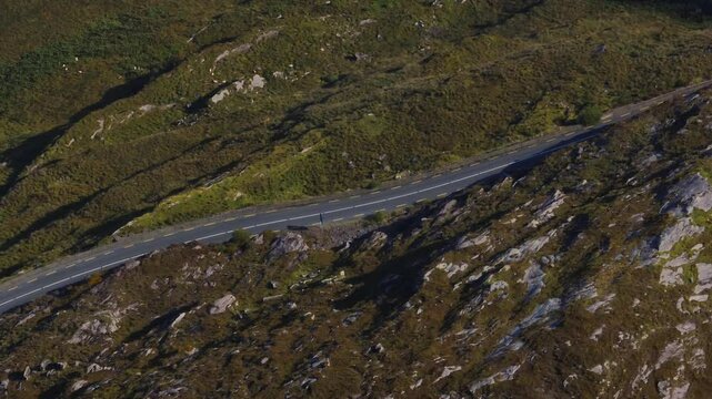 Caha Pass, County Cork, Ireland, September 2024. Drone Bird's Eye View overview follows empty road below white boulders peeking between grass slopes on the border with County Kerry.