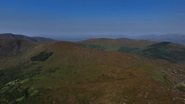 Caha Pass, County Cork, Ireland, September 2024. Drone tracks right revealing a panoramic mountain road crossing the hills into County Kerry on a warm sunny day.