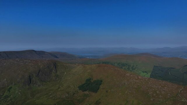 Caha Pass, County Cork, Ireland, September 2024. Drone pulls backwards in panoramic high angle overview descending above rural farmland and forest plantations under a clear blue sky.