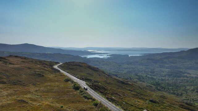 Caha Pass, County Cork, Ireland, September 2024. Drone pushes forward following a car navigating the winding mountain road above rolling hills with the glistening North Atlantic Ocean in the distance.