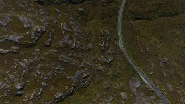 Caha Pass, County Cork, Ireland, September 2024. Drone in Bird's Eye View follows a winding road along mountain edge with exposed rocky cliffs through green grass near the County Kerry border.