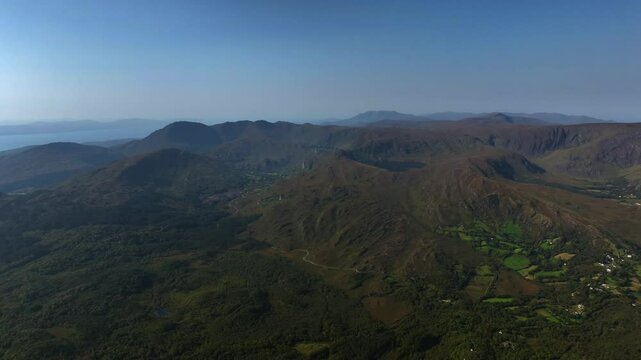 Caha Pass, County Cork, Ireland, September 2024. Drone tracks right in panoramic descent above the rugged mountainous landscape with small fields carved out of planted forests below.