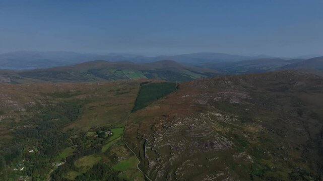 Caha Pass, County Cork, Ireland, September 2024. Drone high angle ascending pullback reveals a panoramic overview with fading mountains on horizon and exposed rock on grassy hillside below.
