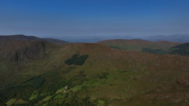 Caha Pass, County Cork, Ireland, September 2024. Drone tracks right above a ridge with vibrant green vegetation and forested regions on the slope with views north to County Kerry in the distance.