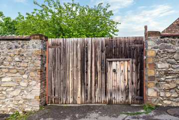 A rustic wooden gate stands tall between aged stone walls, with a smaller door adjacent. Lush green...