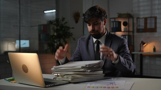 Adult business man manager sitting at desk working on laptop in office overtime in evening, looking through big stack of paper documents, tired expression. - Powered by Adobe