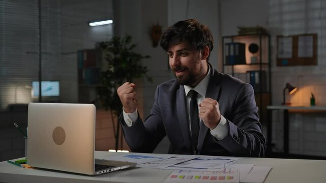 Adult business man manager sitting at desk working on laptop in office overtime in evening, anticipating gets good news, happy expression winning gestures.