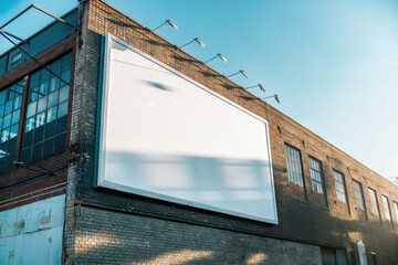 White blank billboard on weathered brick wall with urban building backdrop and clear blue sky, soft sunlight creating an industrial, modern contrast
