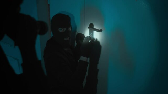 Male burglar in balaclava holding lock pick while trying to open door, unrecognisable crime partner shining flashlight