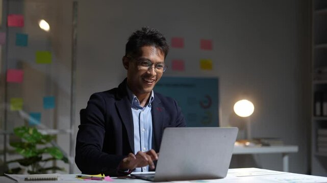 A man is sitting at a desk with a laptop in front of him. He is smiling and he is enjoying his work