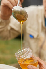 A beekeeper holds a metal spoon from which honey flows into a jar.