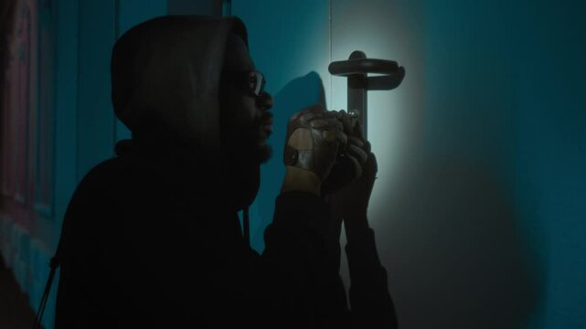 African American male burglar with leather gloves in black sweatshirt opening door using lock pick and nervously looking around at night