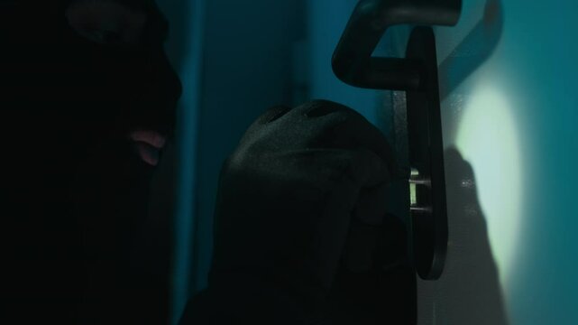 Close-up view of young male criminal with black gloves in balaclava picking lock trying to break into house at night