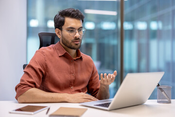 Latin American man businessman participating in online video call on laptop, seated in modern office with casual attire. Engaging in virtual meeting, expressing communication and professionalism.