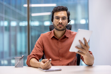 Latin American man businessman in office using digital tablet and headset for video call. Engaged in remote communication, showcasing modern corporate technology and connectivity.