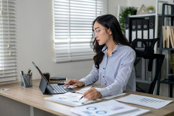 Focused asian businesswoman analyzing financial data on tablet and taking notes in modern office