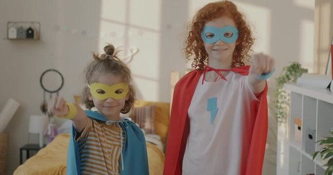 Slow motion portrait of joyful kids siblings wearing costumes masks and capes standing in bedroom raising arms in superhero pose. Childhood and fun concept.
