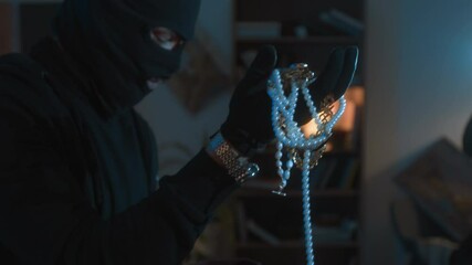Young male robber in balaclava taking jewellery out of box and showing to crime partner while pulling apartment heist