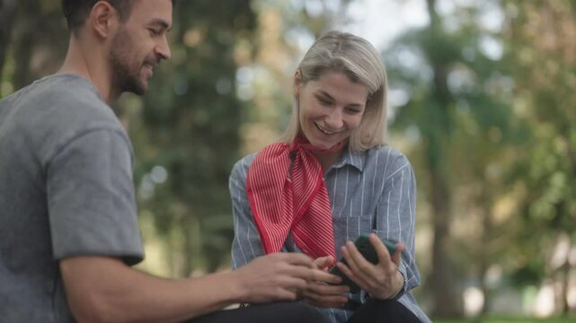 Man and woman looking at mobile phone and smiling. They are sitting in park. Sharing good news or watching something funny together.