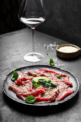 Slices of fresh beef carpaccio with basil and parmesan cheese on a black plate and a glass of red wine vertical photo