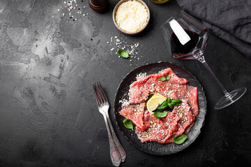 Slices of fresh beef carpaccio with basil and parmesan cheese on a black plate and a glass of red wine on black background top view