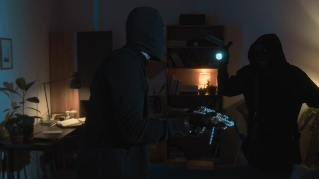 Caucasian thief in balaclava holding jewellery box discussing pearl necklace with Black accomplice with flashlight while holding robbery