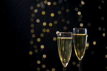 Champagne bubbling in two glasses while blurred golden confetti falling in the background with festive lights, close up shot. New Year party concept.