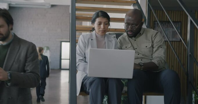 Two businesspeople working with laptop typing and talking sitting on stairs in coworking office. Modern technology and professional cooperation concept.