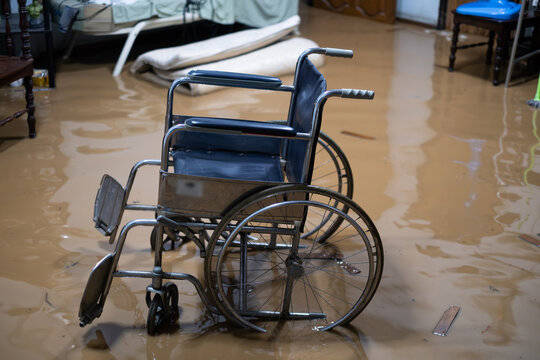 A blue wheelchair is sitting in a flooded room. The chair is upside down and has a broken wheel. The room is filled with water and debris, and there are chairs and a bed in the background