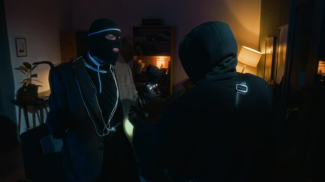 Zoom out view of African American thief shining flashlight at cupboard shelves behind glass doors, his Caucasian crime partner jumping and boasting necklaces and watch during apartment heist