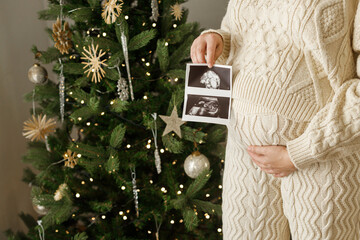 Happy pregnant woman in knitted outfit holding ultrasound scan of baby and hugging belly close up...