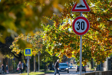 Traffic signs indicating a speed limit of 30 in an autumn street scene with pedestrians