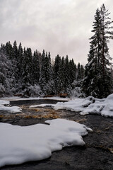 A picturesque snowy river scene with a small dam and rustic wooden structures. The frozen riverbank, pine trees, and overcast sky evoke a cozy, winter wonderland. Ideal for seasonal or rural themes.
