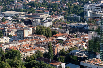 Fototapeta premium A dense urban cityscape combining modern skyscrapers with historic red-roofed buildings surrounded by greenery. Great for themes of city living, architecture, or real estate.