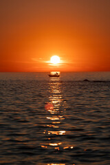 A serene sunset captures the vibrant orange sky and vast ocean, while a silhouetted boat floats gently, creating a peaceful and calming scene on the horizon.