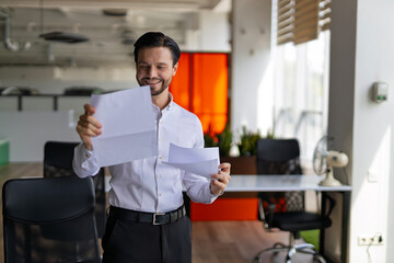 Fototapeta premium A man is standing in a room with a white paper in his hand. He is smiling and looking at the paper. The room has a modern design with a few chairs and a potted plant