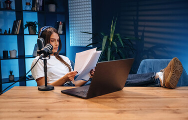 Relaxed, holding paper with text. Woman is hosting radio podcast with microphone and laptop