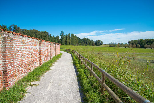 Dirt road on a green meadow in the countryside close to the famous abbey of Morimondo, Italy. Brick wall on the left. Green meadow on the right.