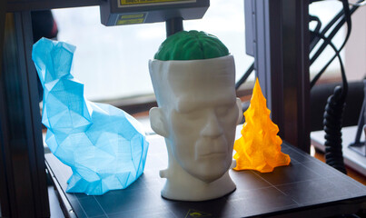 Art models printed on 3D printer on desktop of 3D printer. Models of prototype human head with brain, figured hare and Christmas tree printed on 3D printer from molten plastic of different colors