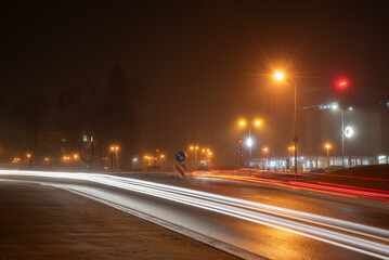  A foggy city street at night with bright car light trails, glowing streetlights, modern buildings, and a hazy urban atmosphere.