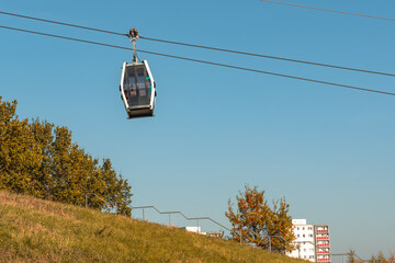 A cable car is suspended in the air above a grassy hill. The sky is clear and blue, and the trees below are bare. The scene is peaceful and serene