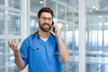 Smiling man doctor in scrubs with stethoscope talking on smartphone, showing effective communication in healthcare setting. Bright hospital environment enhances professional and approachable vibe.