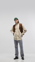 Full-length image of young man man dressed in checked shirt, brown vest, casual jeans paired with green beanie and glasses, posing on studio background. Concept of fashion, youth culture, style, ad