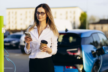 Woman drinking coffee by her electric car while it is charging