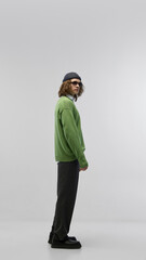 Full-length side view portrait of young confident man in green sweater and gray trousers, sunglasses and hat, posing against light studio background. Concept of fashion, youth culture, urban style, ad